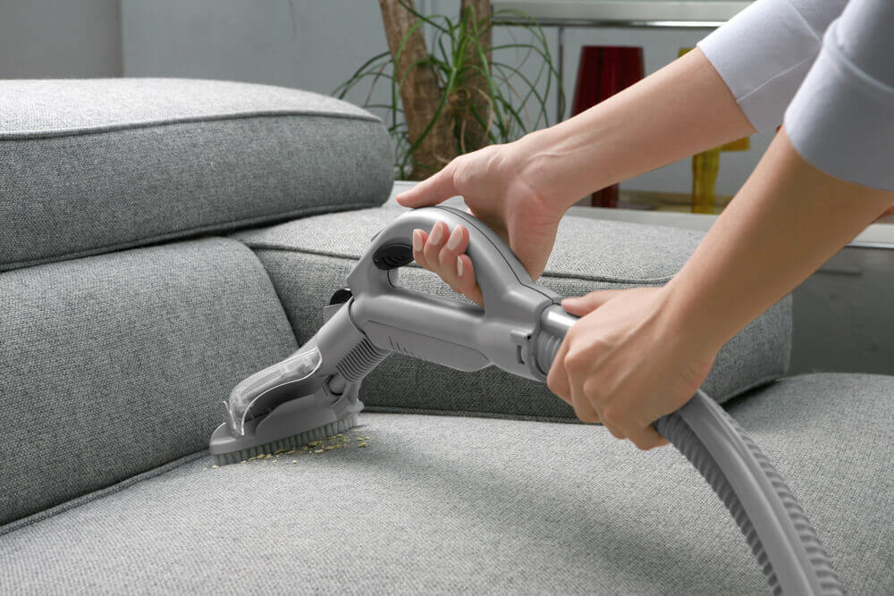 a worker cleaning some crumbs whilst couch cleaning on a grey couch with a course vacuum head 