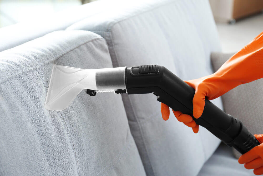 A close up shot of a lounge cleaning wet vacuum head cleaning a light grey couch with a worker with orange protective gloves